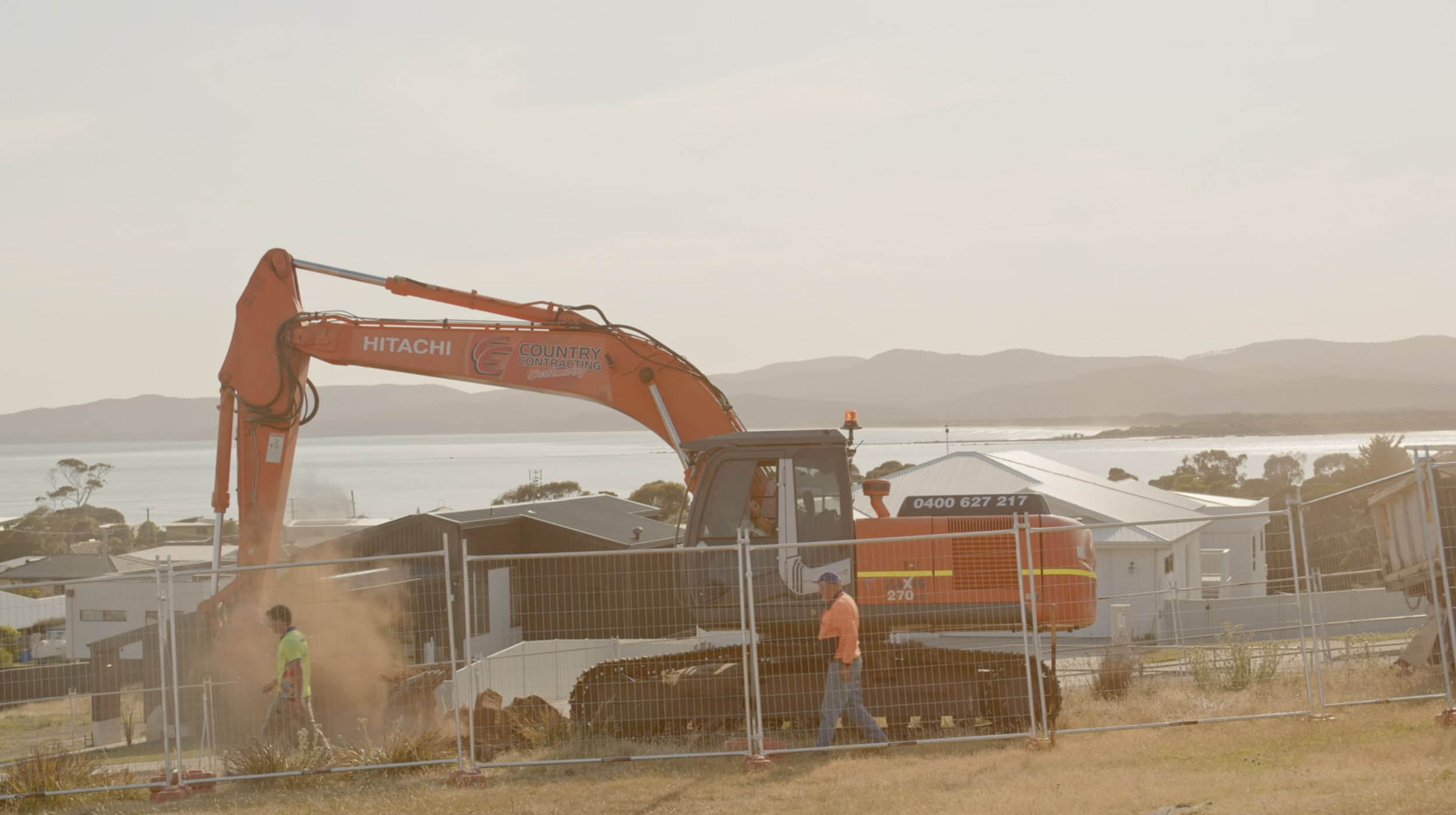 Two people walking past a moving orange excavator.
